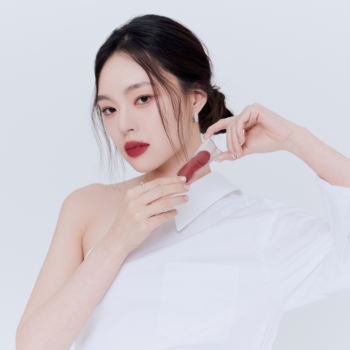 Wegański tint Muzigae Mansion Spicy na ustach modelki; kobieta w białej koszuli trzyma butelkę tintu.