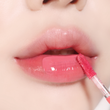 Różowo-koralowy błyszczyk nakładany na pełne, błyszczące usta. Zbliżenie z aplikatorem.