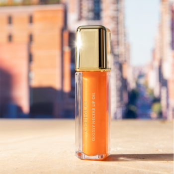 Pomarańczowy olejek do ust Glossy Nectar w przezroczystej butelce ze złotą nakrętką, na tle miasta.