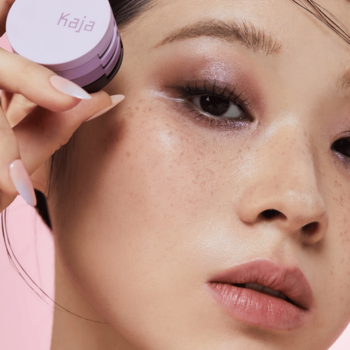 Zbliżenie na kobietę z piegami, trzymającą fioletowy zestaw cieni Kaja Beauty Bento Taro Bubble Tea przy oku. Pastelowe tło.