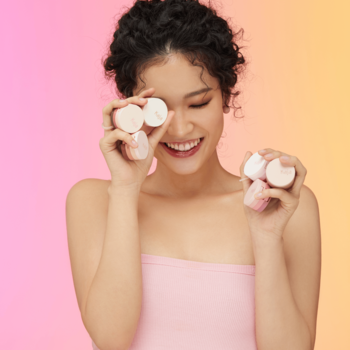 Kobieta z lokami uśmiecha się, trzymając kompaktowe cienie Kaja Beauty Bento Trio Neutral Moment przed oczami.