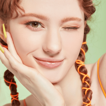 Uśmiechnięta modelka mruga, prezentując ciepły pomarańczowy makijaż oka cieniami Kaja Beauty Bento Peach Madeline.