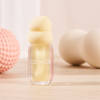 Przezroczysta buteleczka lip oil Catrice z jasnożółtym płynem i warstwową, kremową zakrętką, na jasnym blacie.