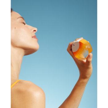 Kobieta w profilu, z zamkniętymi oczami, aplikuje orzeźwiającą mgiełkę do twarzy z pomarańczowo-przezroczystej buteleczki.