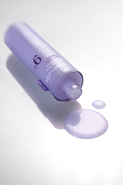 Liliowa butelka toniku rozjaśniającego Numbuzin No.9 leży, z wylaną obok serumową cieczą na białym tle.