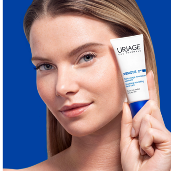 Kobieta trzyma krem Uriage Xemose G+ w białej tubce z niebieskim detalem, na tle jednolitym niebieskim.