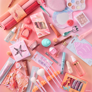 Różnorodne pastelowe kosmetyki: pędzle, cienie, błyszczyki, maseczki i akcesoria z motywem muszli, na różowo-pomarańczowym tle.