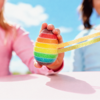 Zbliżenie: Dziecięca dłoń trzyma tęczowe żelki Gumi Yum Surprise w kształcie jajka z paskami żelkowymi.