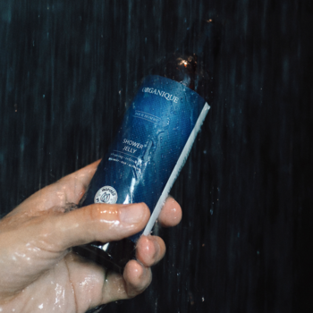Ręka trzyma żel pod prysznic Organique Shower Jelly w ciemnoniebieskiej butelce, w tle woda, widok z bliska.