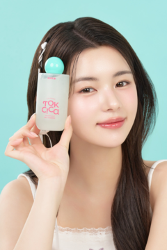 Zbliżenie: kobieta prezentuje EDIT.B Calming Tok Cica Heartleaf Gel Essence, przezroczysta butelka z różowym logo.