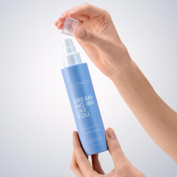 Niebieski spray ICONIQUE DREAMING BIG LIKE YOU do włosów cienkich, otwierany przez dłonie na jasnym tle. Zwiększa objętość.