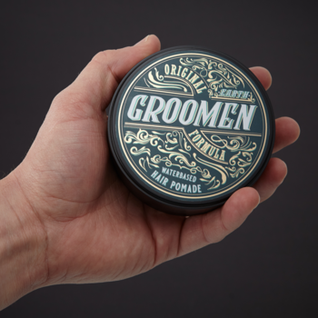 Ręka trzyma okrągłą, ciemną puszkę pomady Groomen EARTH WATERBASED HAIR POMADE z ozdobnym, złotym napisem.