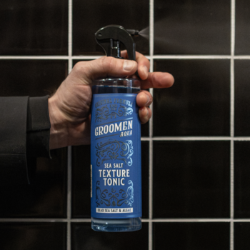 Męska dłoń trzyma niebieski spray Groomen Sea Salt Texture Tonic, tonik do stylizacji włosów.