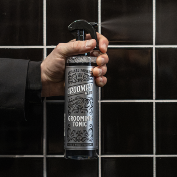 Męska dłoń trzyma czarny tonik Groomen WIND Grooming Tonic z elegancką etykietą, sprayuje, tło czarnych kafelków.