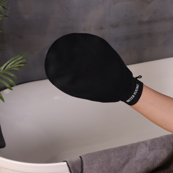 Czarna rękawica peelingująca NOVA Exfoliating Glove SISTER YOUNG z białym logo na dłoni, trzymana nad wanną do złuszczania.