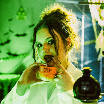 Kobieta w stylizowanym laboratorium o zielonym świetle, kosztuje płyn z kubka. Reklama balsamu Fluff Spooky Spice Latte.