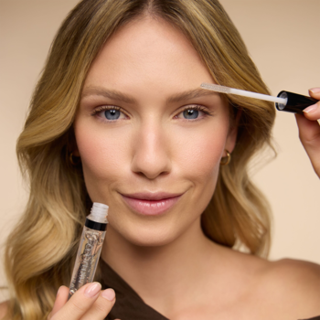 Młoda kobieta aplikuje Clear Lash&Brow Gel, przezroczysty żel do stylizacji brwi, z widoczną butelką w tle.