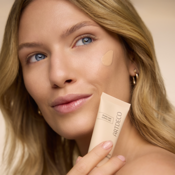 Rozświetlający podkład Artdeco Light Luminous Foundation Warm-Beige Sand na policzku kobiety, w dłoni tubka.
