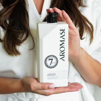 Biała, prostokątna butelka szamponu Aromase Herbal Daily Essential, z czarnym napisem i korkiem, w dłoniach kobiety.
