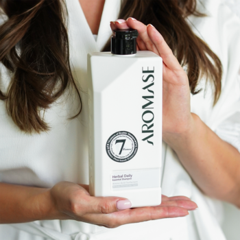 Biała, prostokątna butelka AROMASE Herbal Daily Essential Shampoo z czarnym logo, trzymana pionowo w dłoniach.