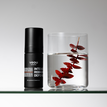 INTENSE REDNESS DEFENSE, krem Veoli do cery naczynkowej w czarnej butelce, obok szklanka z czerwoną rośliną.