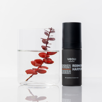 Czarna, matowa butelka Veoli Botanica REDNESS HARMONY obok szklanki z czerwoną rośliną.