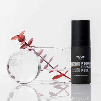Czarny peeling Veoli REDNESS RESCUE PEEL z dozownikiem, obok szklanej dekoracji z czerwoną rośliną.
