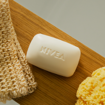 Białe mydło NIVEA Creme Care z wytłoczonym logo, na drewnianej tacce z beżową myjką, widok z góry.