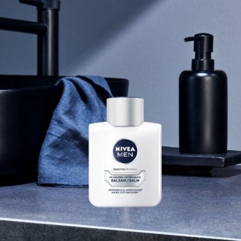 Biała butelka NIVEA MEN Sensitive Regenerujący Balsam z srebrną nakrętką, na ciemnym blacie w łazience.