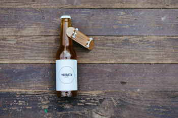 Widok z góry: brązowa butelka mętnej lemoniady Yerba Mate z białą etykietą, na niej mini deskorolka, na drewnie.