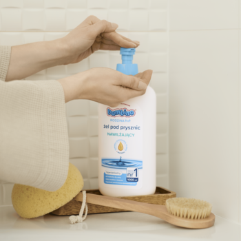 Ręce naciskają pompkę białego nawilżającego żelu pod prysznic Bambino Rodzina 1000 ml na drewnianej tacy.