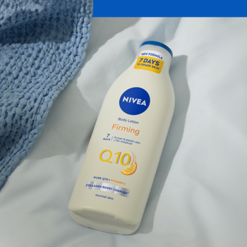 Biała butelka ujędrniającego balsamu Nivea Q10 + Witamina C na jasnym prześcieradle, widok pod kątem.