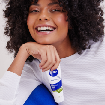 Uśmiechnięta kobieta z kręconymi włosami trzyma biały krem do rąk NIVEA Moisture Care z oliwą z oliwek.
