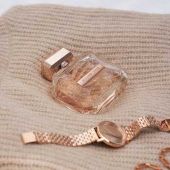 Kwiatowo-drzewna woda perfumowana Jennifer Lopez Promise w szklanej butelce i zegarek z różowego złota na beżowej dzianinie.