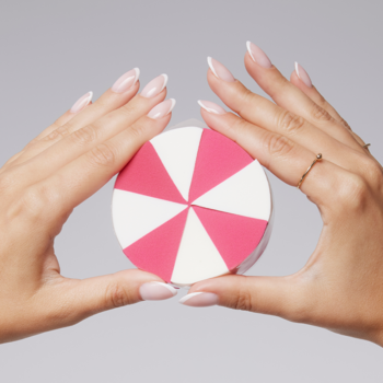 Ręce trzymają okrągłą gąbkę do makijażu z różowo-białymi trójkątnymi wzorami, na szarym tle.