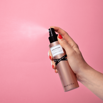 Dłoń z czerwonymi paznokciami rozpylająca mgiełkę Miss So...? Love Potion z różowo-złotej butelki.