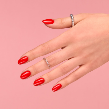 Dłoń z lakierem hybrydowym Semilac 039 Sexy Redy, intensywna czerwień, błyszczące paznokcie, srebrne pierścionki, różowe tło.