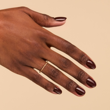 Ciemnobrązowy lakier do paznokci o ciepłym, pełnym kryciu, na dłoni z delikatnym złotym pierścionkiem.