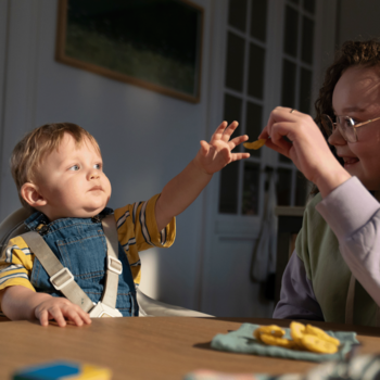 Niemowlę w niebieskich ogrodniczkach wyciąga rękę po kawałek owocu podawany przez dorosłego, w kontekście musów BoboVita.