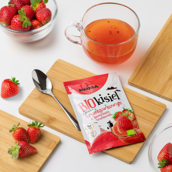 Czerwona saszetka BIO kisiel truskawkowy z owocami, obok szklanka kisielu, świeże truskawki na deskach.