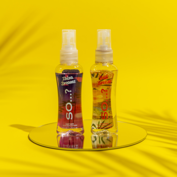 Dwie mgiełki So...? Escapes Ibiza Dreams, w przezroczystych butelkach z barwnymi etykietami, na lustrze na żółtym tle.