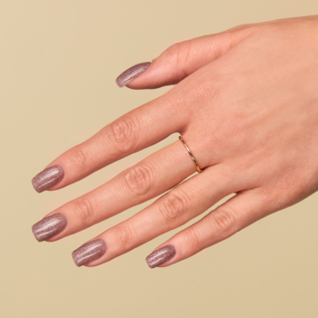 Dłoń z hybrydowym lakierem w jasnym brązie z fioletem i srebrnym brokatem, złoty pierścionek. Widok z góry.