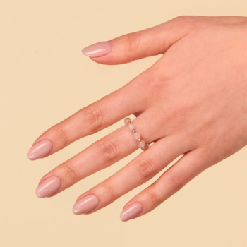 Ciepły beżowy lakier hybrydowy nude z brokatowym pyłkiem na paznokciach. Dłoń z delikatnym pierścionkiem.