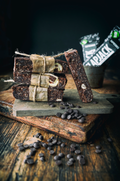 Wegańskie batony owsiane z nerkowcami i czekoladą na drewnianej desce. Trzy owinięte sznurkiem, jeden odkryty. Wokół czipsy.