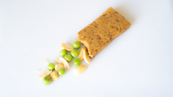 Ugryziony wegański baton proteinowy z nerkowców na białym tle, obok zielony groszek i jasne płatki.