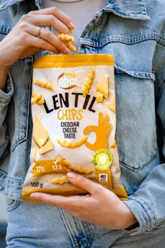 Beżowa paczka chipsów soczewicowych Ohoi Cheddar Cheese Taste z białym napisem 'Lentil Chips', trzymana przez osobę w jeansowej kurtce.