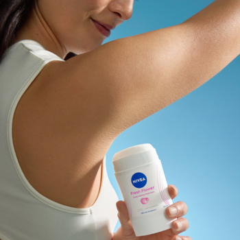 Kobieta aplikuje biały antyperspirant w sztyfcie NIVEA Fresh Flower pod pachę, na jasnoniebieskim tle.