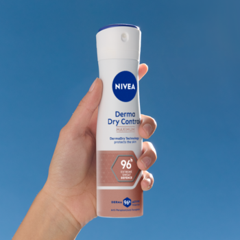 Biały antyperspirant NIVEA Derma Dry Control 96h w sprayu, trzymany przez dłoń na tle niebieskiego nieba.
