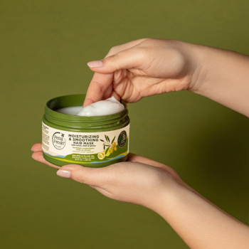 Ręce kobiety nabierają palcami nawilżającą maskę do włosów z zielonego słoiczka na oliwkowym tle.