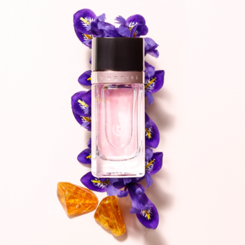 Różowa butelka perfum Bugatti Eleganza, z czarną nakrętką, leżąca na fioletowych kwiatach irysa i bursztynie.